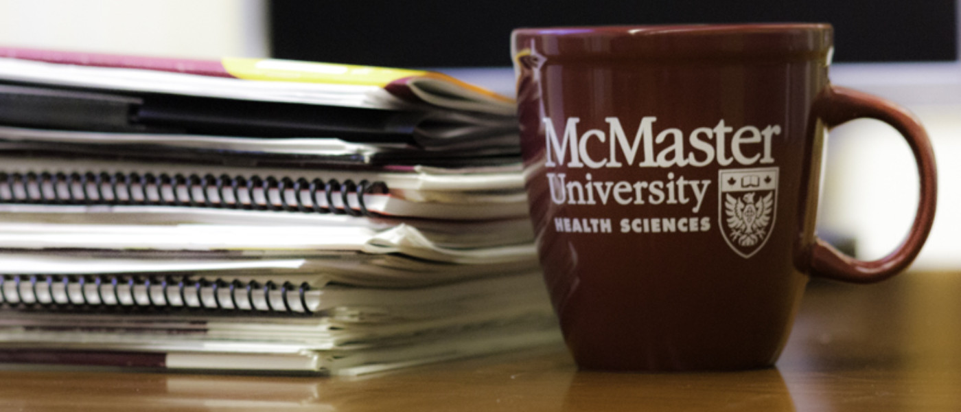 A McMaster University Health Sciences coffee cup beside a stack of books.
